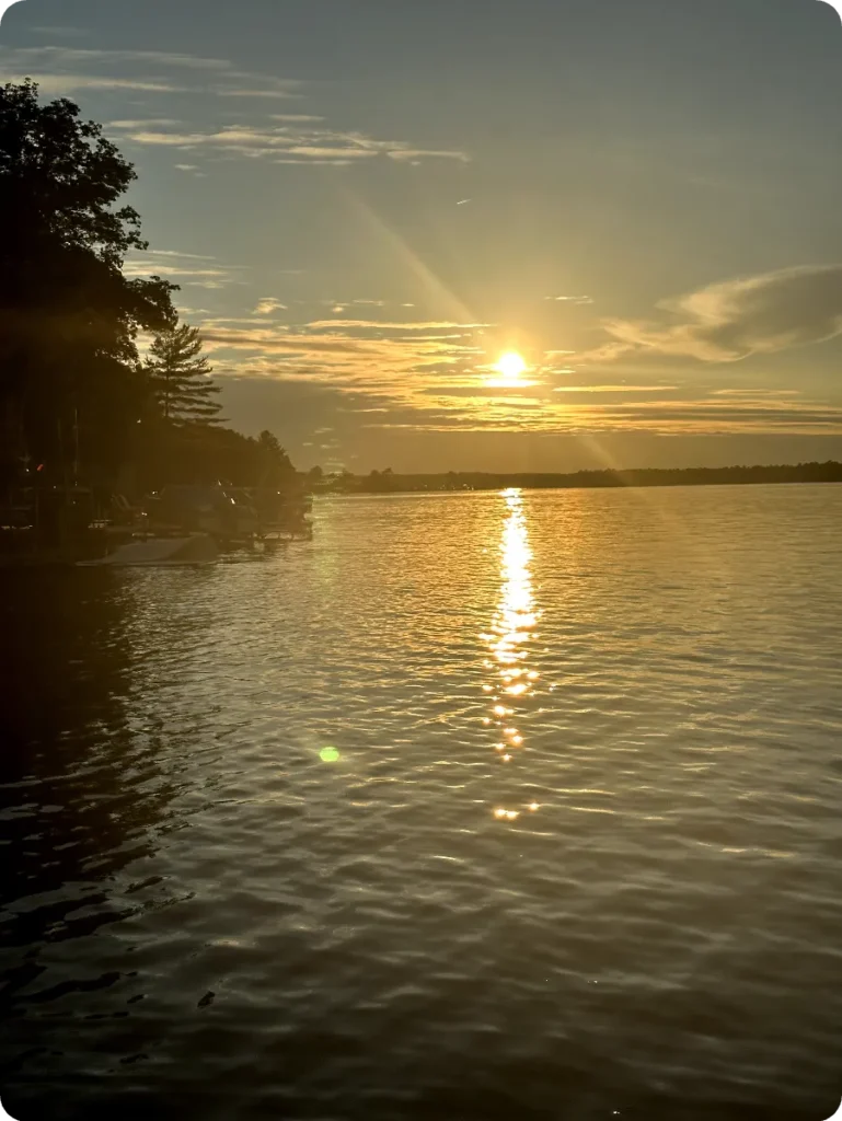 A sunset over a calm lake with a silhouetted tree line on the left and the sun reflecting on the water.